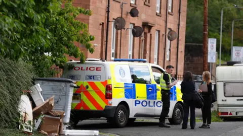 A police officer stands with two women in front of a police van in the middle of a street. 
