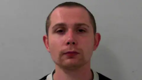 Mugshot of a man with a short goatee and a buzzcut. He has a neutral expression. He is wearing a grey jumper.