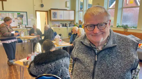 A man with short white hair, glasses, a grey bodywarmer and a green and white checked shirt is standing in a church hall. Behind him, a woman is stood reading from a piece of paper. There are another three women and two men sat at trestle tables. The walls are cream and the floor is wooden. There are cupboards, a large noticeboard, a clock and a crucifix on the far wall and four windows on the right wall.