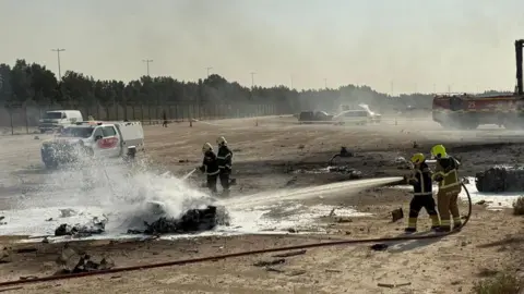 Dubai Media Office Firefighters work at the scene of a plane crash, water spraying onto a burnt piece of metal on the ground, it is in an open stretch of land with cars and a fence visible in the background.