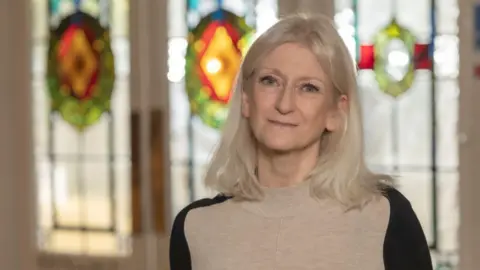 North Yorkshire Council A middle-aged woman with short light blonde hair and a beige jumper with black sleeves stands in front of a stained glass window,