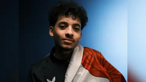 A young man with black hair is looking at the camera, he is wearing a black jacket and has the Welsh flag draped on his left shoulder. The background is a wall that is a mix of light and dark blue.