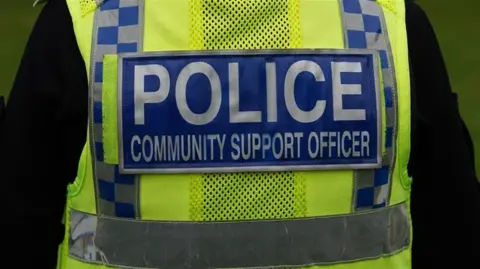 Picture of the back of a Police Community Support Officer in uniform with a hi vis police jacket on. The jacket has white writing on a blue background with the word police in bold. 