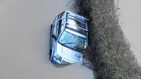 A silver Land Rover stuck in flood water. It is slightly tipped to the side and water is coming up over one of its headlights. 