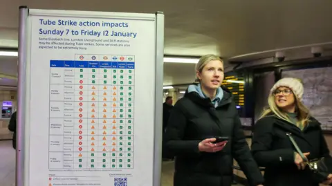 An image of a poster with information about the Tube strike action. Two women walk past the sign wearing long black puffer jackets and one wearing a wooly hat. 