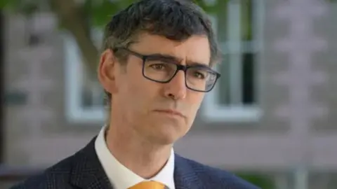 BBC Deputy Montford Tadier is wearing a blue blazer and a yellow tie. He has glasses and brown hair