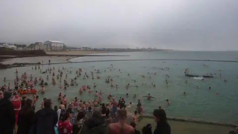 Dozens of people - some of whom are wearing fancy dress - go for a swim in a lido in Jersey as part of a Christmas Day Swim event. The weather is cloudy. A number of people are stood on the side of the lido watching people in the water.