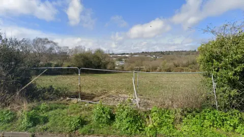 A view of a grass field through a gap in the hedgerow looking out over Hastings. There are three temporary metal fences blocking the entrance. The sky is blue and cloudy