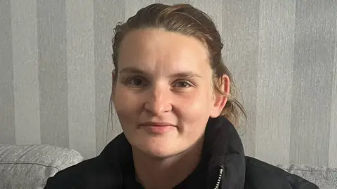 BBC A woman with brown hair tied back wearing a black coat, directly facing the camera, with striped grey wallpaper in the background