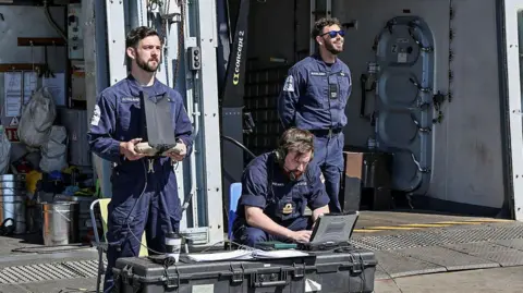 LPhot Robert Oates Three men, two standing one sitting, operate drones from a frigate deck wearing Royal Navy boiler suits 
