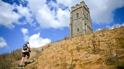 Finnbarr Webster/Getty Images A general view of the dried land at Glastonbury Tor. A man wearing a back pack shorts and a shirt is walking up a set of steps to the left. He has his back to the camera.