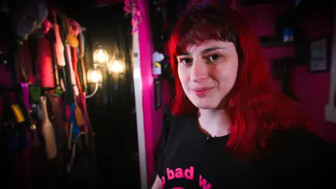BBC Porcelain Victoria is smiling at the camera. She has long hair, dyed bright red hair. She is wearing a black T-shirt with bright pink text on it. On the left hand side there is a collection of straps and paddles, arranged on hooks. The room is dark.