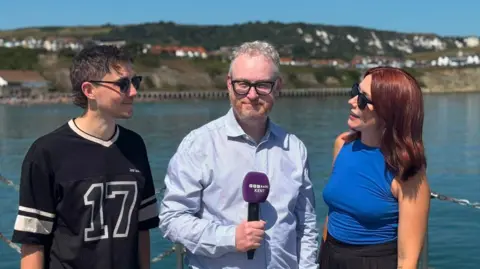 BBC Sam and Bec stand with BBC Radio Kent presenter Dominic King beside a waterfront railing on a sunny day, with water, chalk cliffs, hillside homes and seafront buildings in the background.