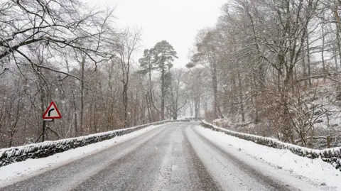 The A591 in Grasmere is a two lane rural road, with stone wall barriers on either side and a red sign indicating a sharp bend ahead. The road is dusted with snow, as are the tall trees bordering the road. Snow is falling.