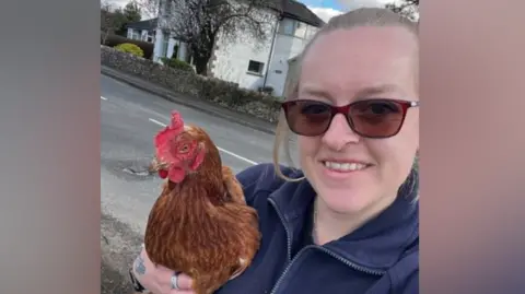 Heather Shepherd Heather Shepherd stands in a road holding a chicken. She is wearing brown sunglasses and her blonde hair is tied back. She is wearing a navy fleece.