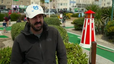A man in a black jacket and a white cap. A busy crazy golf course featuring a stripy lighthouse is behind him.