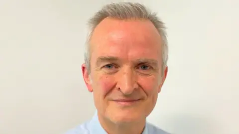 West Suffolk Hospital Dr Ewen Cameron is smiling at the camera. He has grey hair and is wearing a light blue shirt and standing in front of an off-white coloured wall.