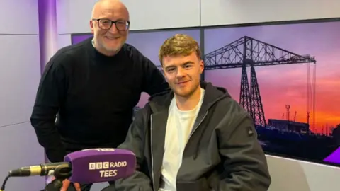 Tommy Conway in the Radio Tees studio with a purple microphone in front of him. He is smiling and looking into a camera. There is a brightly lit background image of the transporter bridge behind him. 