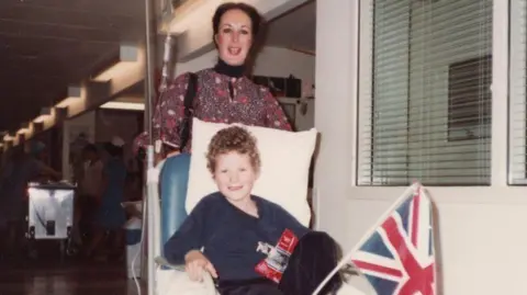 PA Media Antonya Cooper stands behind her young son Hamish who sits in a hospital chair.