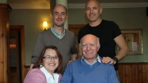 Daniel Hicks Two men stand in front of a woman and older man who sit on chairs. They are all smiling at the camera. The men are bald and the woman has short brown hair. The man at the top left wears a grey jumper with a blue shirt underneath. The man on the top right wears a black T-shirt. The man below wears a blue jumper and shirt. The woman wears a pink cardigan and a white blouse.