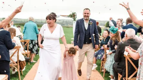 James Kaminski A happy bride and groom walk down the aisle holding the hand of a young bridesmaid in a pink dress.
