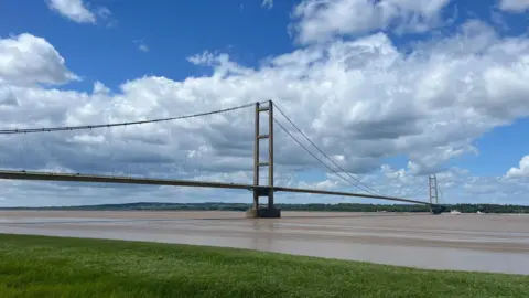 Local Democracy Reporting Service The Humber Bridge, stretching across the water on a sunny day with nimbus clouds overhead.