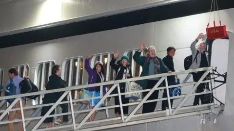 PA Media Seven passengers boarding the ship from a platform. Four of them are waving.