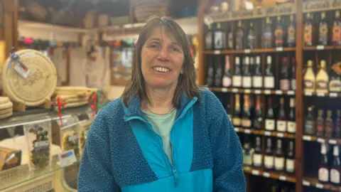 A woman with short brown hair. She is wearing a light green top and blue fleece. Behind her to the right is a fridge and behind her to the left are shelves with wine.
