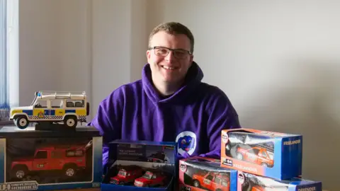 Valart Ltd A smiling man sat behind his collectibles of Land Rover models.