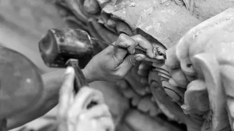 Getty Images A close-up black-and-white image shows hands using a small chisel and hammer to carve fine details into a piece of stone. One hand holds the chisel while the other grips a rounded mallet. Small chips and dust are visible on the stone surface, which shows intricate shapes and textures.