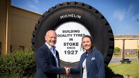 Two people stand in front of a large Michelin tyre, shaking hands. The middle of the large tyre has black text on a white background saying "building quality tyres in Stoke since 1927."