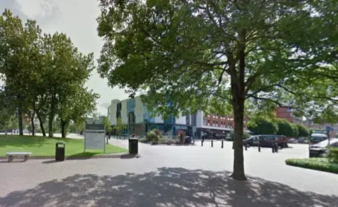 Aston University campus. There is a sign with directions, as well as trees and buildings with grass areas, some pillars and shaded areas by the trees. 