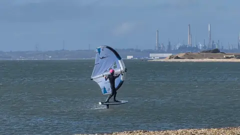 stacey johns A windsurfer crosses the water. Factories are visible on the coast in the distance.