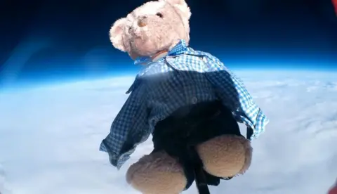 mall, brown bear, wearing school uniform, navy blue shorts and a blue and white checked shirt flying attached to a weather balloon above clouds