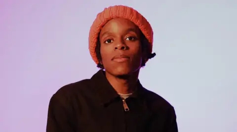 Johnathon Williams A man with a red knitted hat is looking at the camera, and has black hair underneath it. The backdrop is a light purple which fades into a light blue/white. He is wearing a black sweater