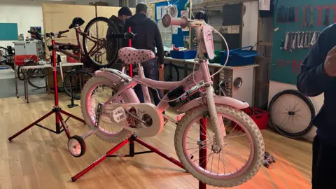 Grace Wood/BBC A pink children's bike hanging on a red stand. In the background two men huddle around a toolkit.