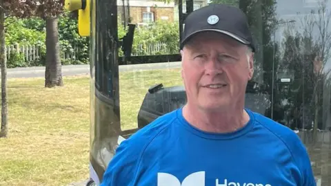 A man standing in front of a bus wearing a blue T-shirt which reads HAVNS HOSPICES. He wears a baseball cap. There is grass and trees to the left of the bus.