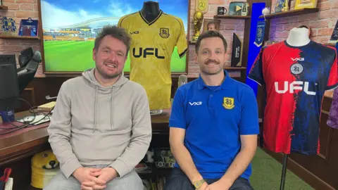 Warren Muggleton/BBC Two men sat in an office, smiling at the camera with lots of football shirts around them.