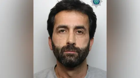 A man with black hair and a black beard wearing a grey tshirt, standing against a grey background