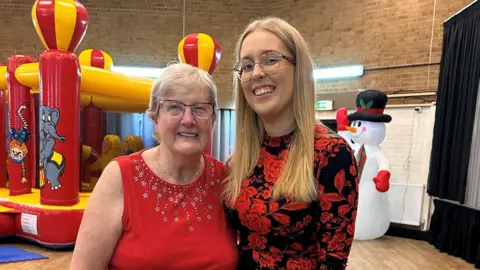 Supplied Shannon has blonde hair, wears a red floral-patterned dress, and is arm and arm with her nan Babs, who wears a red top. Babs has glasses and fair hair. They are inside a hall, with a red and yellow bouncy castle behind them.