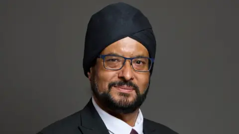 A headshot of a man in a turban on a grey background.