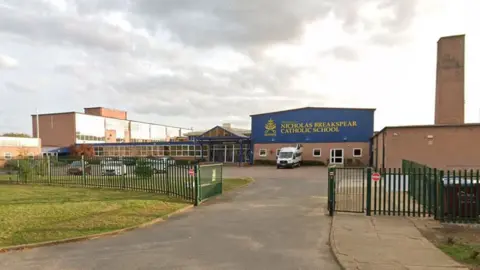 A school building seen from outside the school gates. It has green metal fences. The main building is partly blue with yellow writing, reading "Nicholas Breakspear". The rest of the building is largely made up of windows.