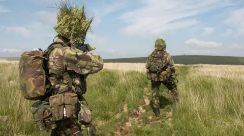 Two camouflaged British soldiers make their way through an open field towards a helicopter on the horizon.  