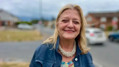 Martin Giles/BBC Julia Ewart is standing on a street with homes, cars and a green in the background. She is wearing a blue denim jacket, a coloured top and large taupe coloured necklace. 