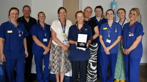 Somerset NHS Foundation Trust A group of midwifery staff in uniform, smiling to camera and surrounding Pip who is holding a certificate