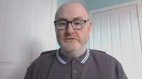 A man sits in front of a white panelled wall. He is wearing a polo shirt buttoned up to the neck. It is purple. He is bald, unshaven and is wearing glasses