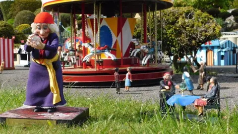 Bekonscot A view of a fairground in a model village. A larger puppet of a fortune teller holding a crystal ball is stood close to where a miniature version of the same character is giving a reading as part of the model village.