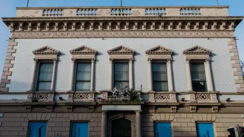 A beige and white square building with five windows on the first floor and a crest above the door. Each window has a pillar on either side, and there are a further four windows on the ground floor.