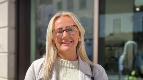 Lauren Handscomb has long light-coloured hair. She stands outdoors in bright sunlight, facing the camera. She wears patterned glasses, a light knit top, and a beige coat, with small gold earrings visible. Behind her is a building facade with a large window and reflections.