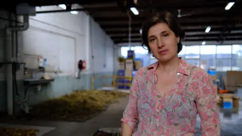 BBC/Katy Watson A brunette woman with short hair wearing a pink paisley top looks at the camera, dried flower petals in the background behind her